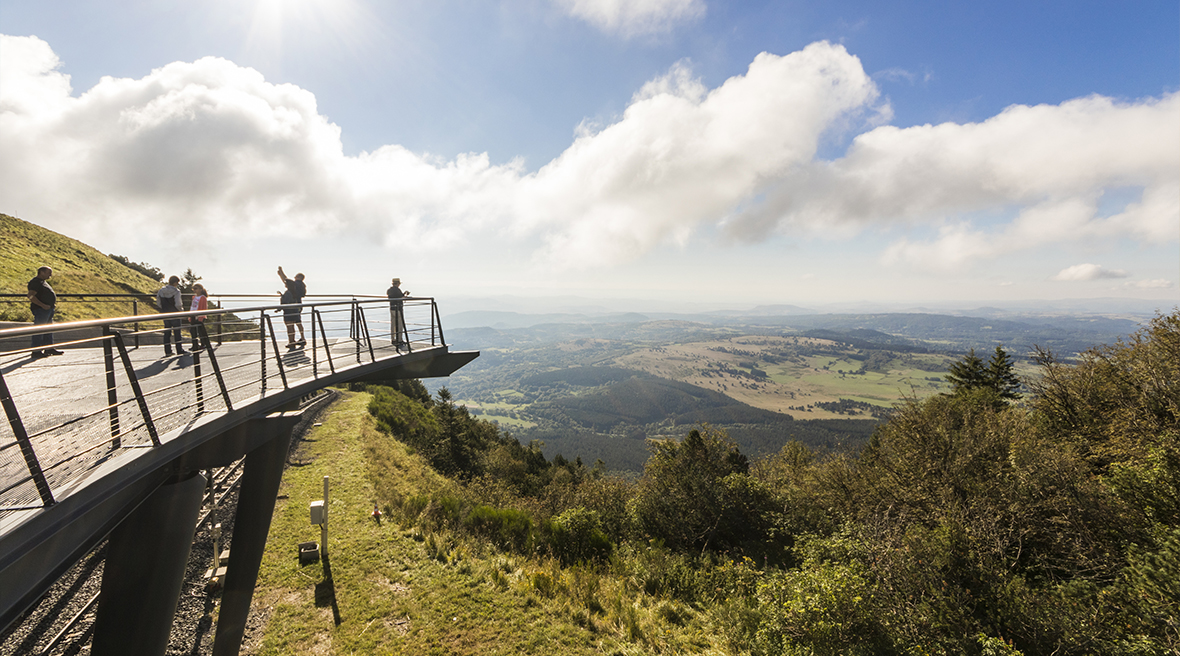 People standing on a walkway on the side of a mountainous volcano with big blue skies and greenery spreading below